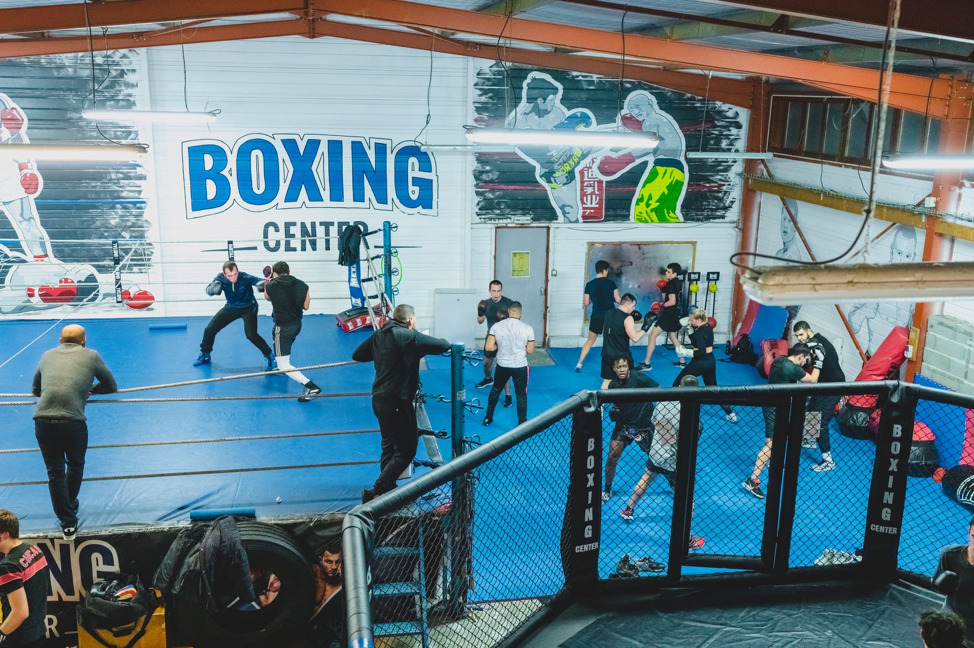 La salle vue depuis l’étage : le ring bleu à gauche, l’octogone grillagé à droite, et « Boxing Center » peint en bleu sur le mur blanc.