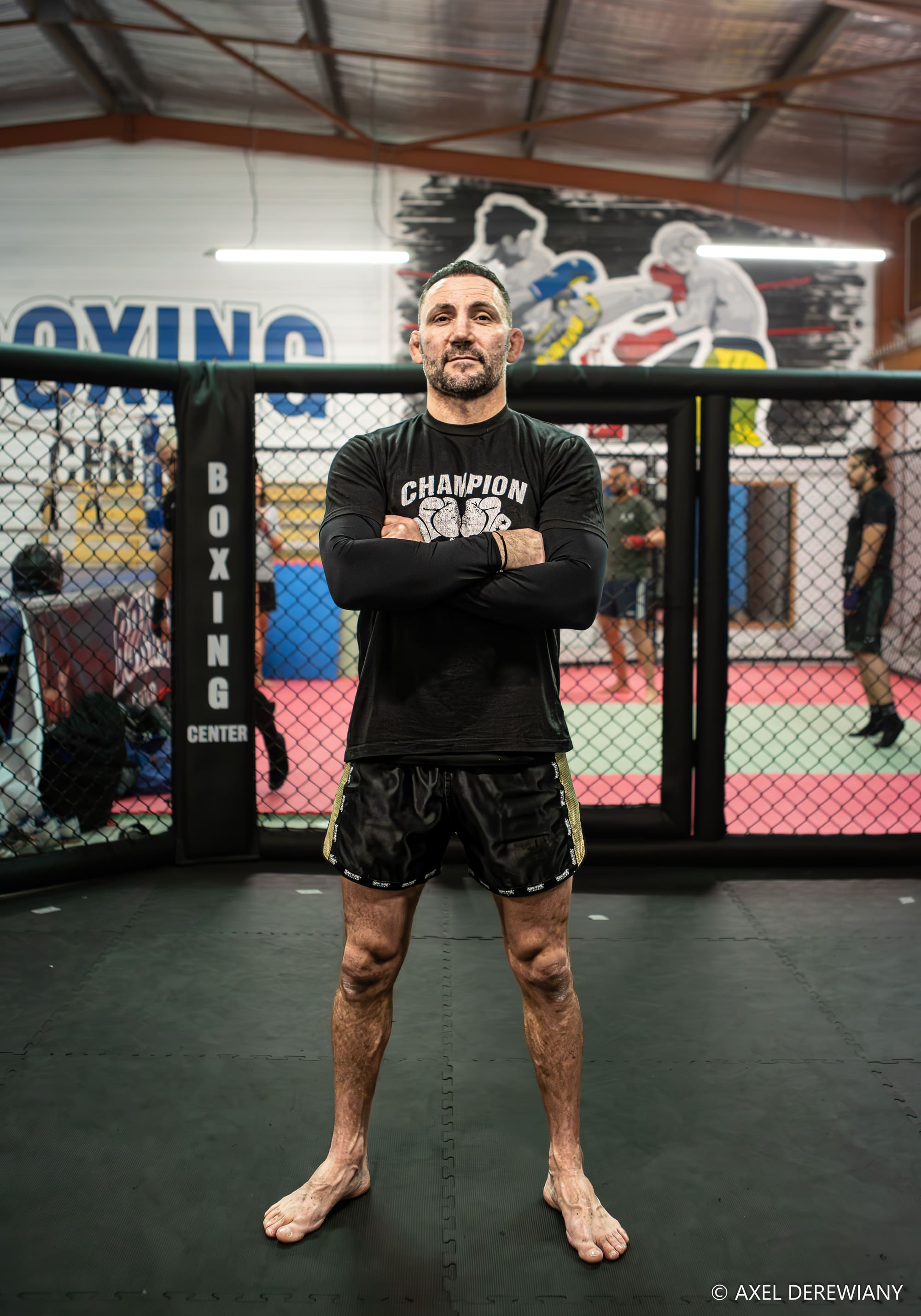Un coach en t-shirt « Champion », bras croisés, debout devant le grillage de la cage.