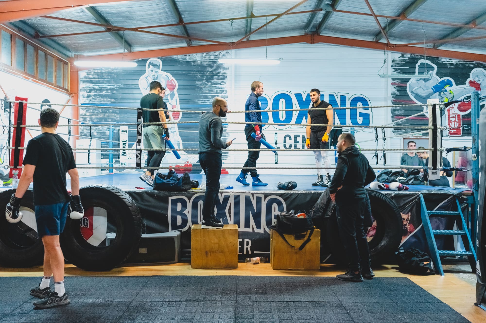Un cours sur le ring : plusieurs pratiquants debout entre les cordes, d’autres autour, gants aux mains.