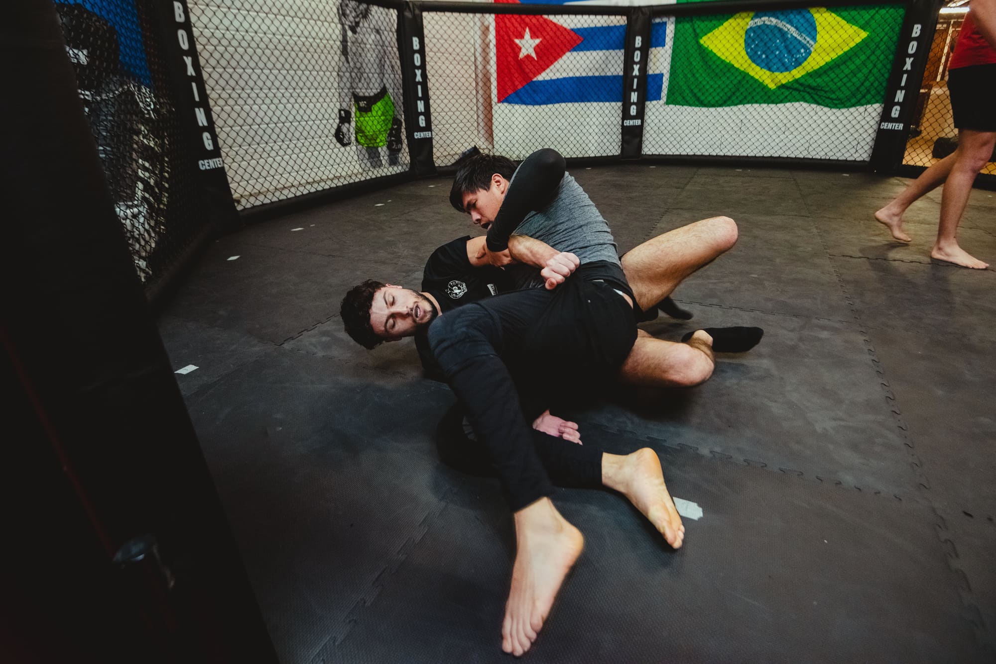 Deux pratiquants au sol en grappling à l’intérieur de la cage, les drapeaux accrochés au grillage derrière eux.